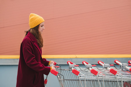 Young beautiful woman in a burgundy coat and yellow hat walks on the roof of supermarket parking lot and takes trolley.の写真素材