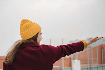 Young beautiful woman in burgundy coat and yellow hat walks along the roof of the parking lot....