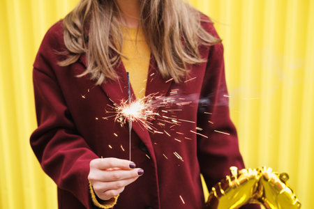 Unknown woman in burgundy coat and holding a burning sparkler in her hands against the background of bright yellow wall.の写真素材