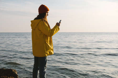 Young beautiful woman in bright yellow coat stands on the seashore or ocean and make a photo on smartphone. Concept of freedom, thoughtfulness, mindfulness.の写真素材