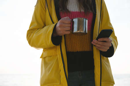 Young beautiful woman in bright yellow coat stands on the seashore or ocean with cup of tea, coffee...