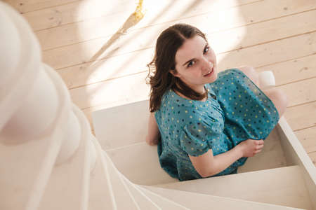 Young beautiful woman in a blue dress with polka dots sits on the stairs and looks at the camera, smiling.の写真素材