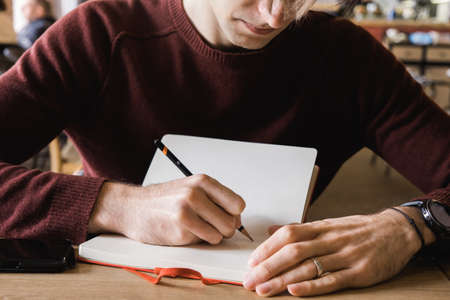 A young man sits in cafe, drinks coffee and makes notes and sketches in notebook.