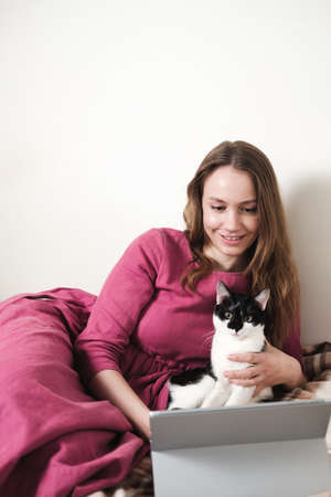 Young millennial woman in pink dress with long hair using a tablet and watcing movies with her black and white tuxedo cat at home.の写真素材