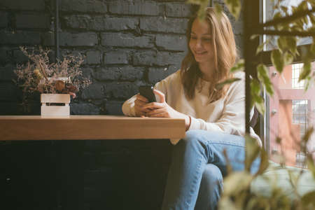 Millenial generation young girl sitting in the cafe at a table, looking at smart phone and smiles.の写真素材