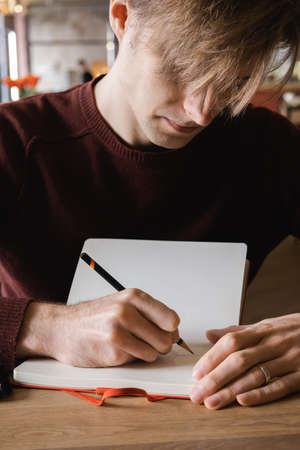 A young man sits in cafe, drinks coffee and makes notes and sketches in notebook.の写真素材