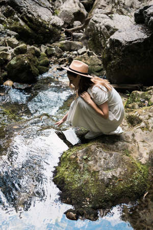 Beautiful woman in white long dress and hat is sitting by the river and smiling. Travel concept.の写真素材