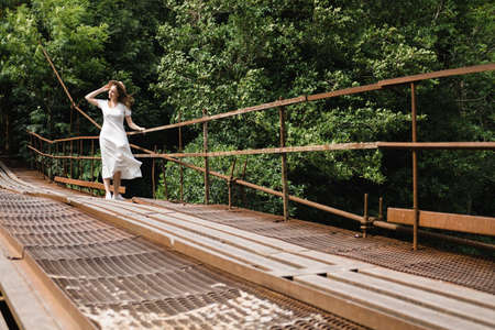 Beautiful young millennial woman in white long dress and hat is walking on the bridge and smiling. Travel concept.の写真素材