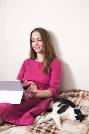 Young millennial woman in pink dress with long hair drink wine and using a tablet with her black and white tuxedo cat at home.の写真素材