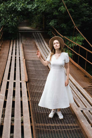 Beautiful young millennial woman in white long dress and hat is walking on the bridge and smiling. Travel concept.の写真素材