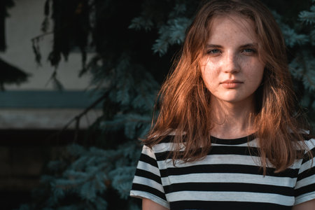 Portrait of young lovely woman with blue eyes and freckles in striped T-shirt and jeans against background of green foliage.の写真素材