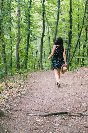 Young millennial woman in dress and beige hat stands in forest happy and relaxed, enjoys nature and...