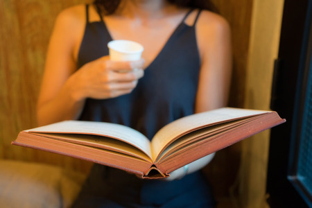 Young girl is sitting on a sofa and reading a book while holding a paper cup