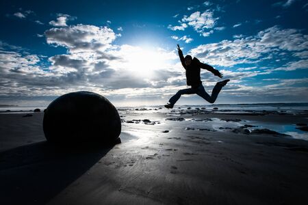 backlight shot of jumping happy young man at boulders beach in New Zealand. Man in the right side, one of the boulders on the left.の写真素材