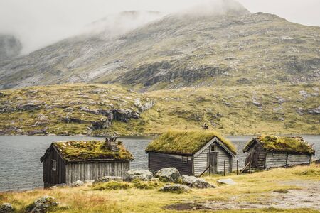 three typical norwegian wooden houses with gras on top, lake and mountains in backgroundの写真素材