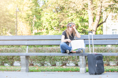 confounded, lost or thinking tourist woman looking at map for right way, sitting on the bench in the park,  luggage on the streetの写真素材