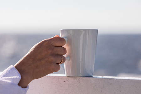 White mug in a womans hand on the terrace edge, romantic mood with morning coffee. Blurred sea in backgroundの写真素材