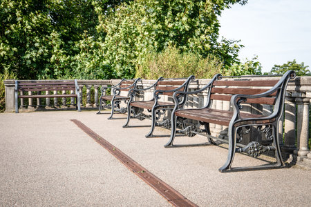 A row of classic wooden benches with ornate black iron frames stands neatly aligned beside a stone...