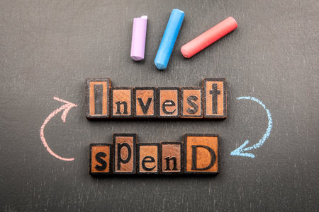 Words invest and spend made from vintage wooden letterpress blocks with chalk arrows on blackboardの写真素材