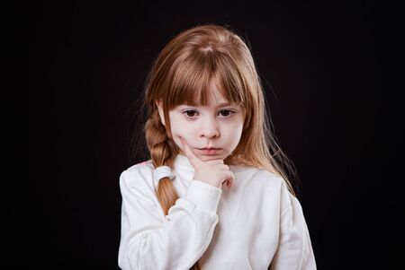 little blonde girl looking thoughtfully down finger on cheek on black backgroundの写真素材