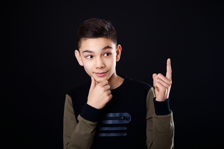 The boy thinks. Thoughts. The guy shows a finger up, an idea. Place for text. Black background, studio, isolated.の写真素材