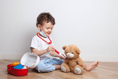 funny little boy in a medical mask, a toddler in a doctor's suit with a stethoscope makes an injection to a teddy bear. game in the profession. vaccination. teaching children to medical procedures.の写真素材