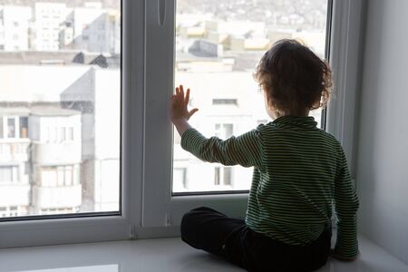 a boy dreams of going out sitting on quarantine at home by the window. the child sits on a...
