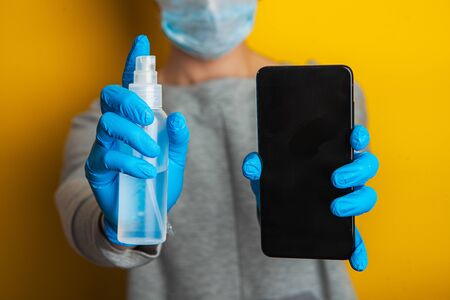 disinfection of the phone. A woman in a medical mask and rubber gloves holds a telephone and a spray disinfector in her hands. protection of electronic devices from coronavirus transmission.の写真素材
