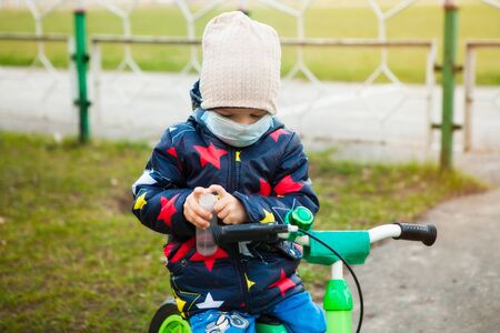 child on a walk in a city park in a medical mask on a bicycle. Hand treatment with antiseptic....