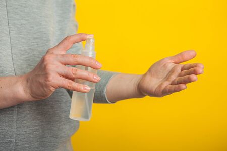 a woman in a medical mask disinfects her hands with a spray sanitizer on a yellow background,...