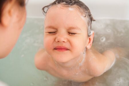 Mom washes her baby's head in a bath with water. Flushing shower shampoo foam. Shampooing without...