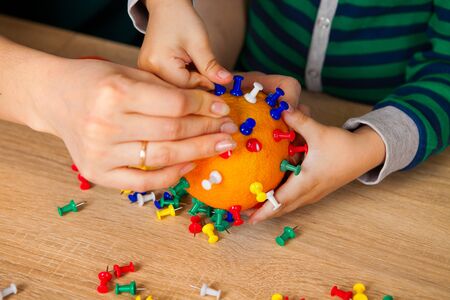 stay at home. Mom with a child makes a dummy of a coronavirus from orange and buttons. explaining to...
