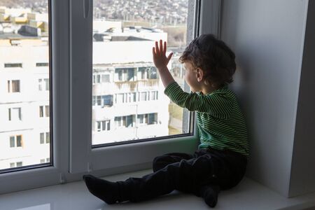 cute curly baby looks out the window into the street while sitting on the windowsill during quarantine of the coronavirus.の写真素材
