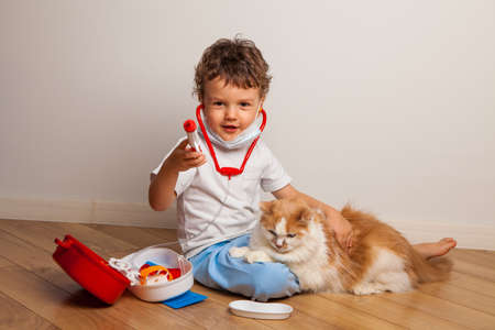 funny curly kid in a medical mask and glasses with a stethoscope on his neck plays a doctor with a cat. The child wants to inject the cat with a toy syringe.の写真素材
