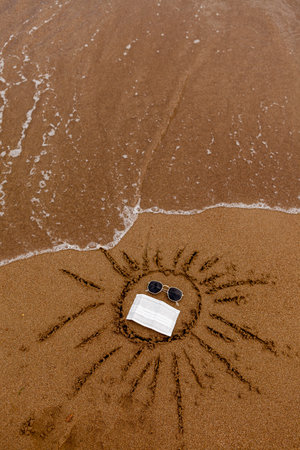 Drawing sun sunglasses and a medical mask on the sand of the beach. Rest at sea and coast during the pandemicの写真素材