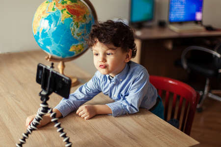 A child blogger shoots a video on a smartphone on a tripod. The boy poses on a smartphone camera, shoots a vlog, next to the globe. Funny cute kid.の写真素材