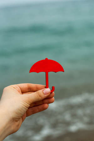 the hand holds a miniature red umbrella against the background of the sea. Symbol the figure of an umbrella on the seashore. Protection from bad weather. Climate and bad weather on summer sea holidaysの写真素材