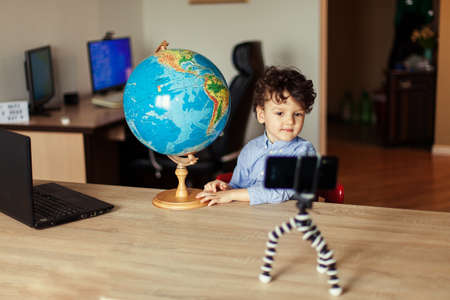 A child blogger shoots a video on a smartphone on a tripod. The boy poses on a smartphone camera, shoots a vlog, next to the globe. Funny cute kid.の写真素材
