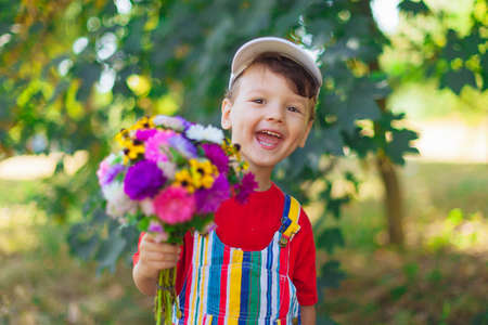 laughing boy with a bouquet of flowers. A child with a bouquet of wild flowers for mom, a gift, a surprise with their own hands. Emotional kid with flowers in bright clothes in natureの写真素材