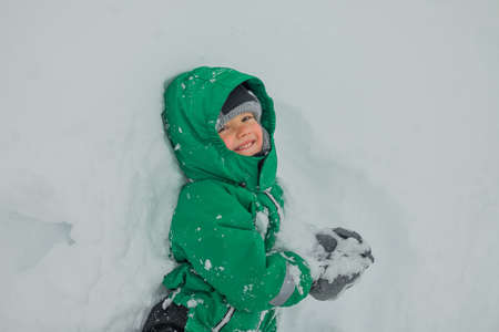 A happy laughing child lies in the snow in a snowdrift. Snowball games, winter games outside. A preschooler in a green jumpsuit in the snow.の写真素材