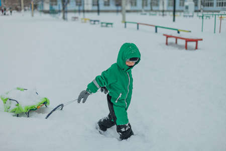 a lifestyle portrait of a toddler dragging a sled through the snow. Winter Games. A happy child in a green jumpsuit carries a sleigh through snowdriftsの写真素材