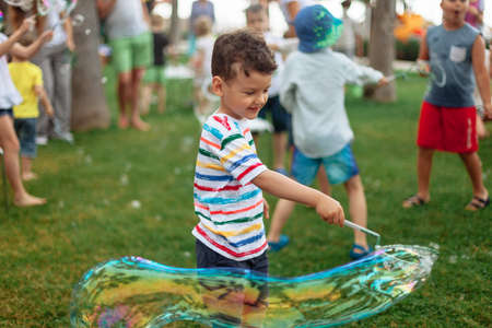 the child inflates large soap bubbles. Street animation program, happy child on a green lawn. Summer vacation. A boy in bright striped clothes with soap bubbles. Large portrait, horizontalの写真素材