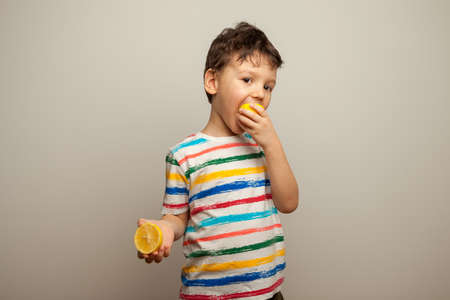 little boy eating lemon, sour taste, makes grimace, facial emotions negative, in white striped T-shirt, isolated white background, copy space. High quality photoの写真素材