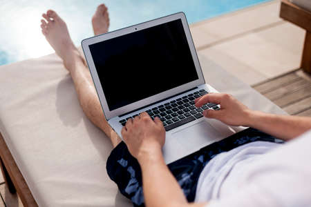 a man works at the computer lying on a sun lounger by the pool. Close-up of hands on a laptop without a face.の写真素材