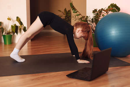 child, the girl sits in front of the laptop on the sports mat and repeats the exercises online in stretching. online home workouts for kidsの写真素材