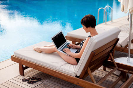 a woman in a swimsuit with a laptop lies on a sun lounger by the pool and looks at the monitor screen. work online. swimming pool on site.の写真素材