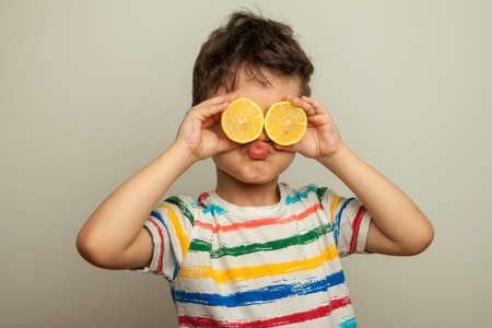 a funny emotional boy, a child with yellow fresh lemons in his hands closes his eyes with lemons and makes a face. A boy with citrus on his eyes instead of binoculars. Vitamins, healthy nutritionの写真素材