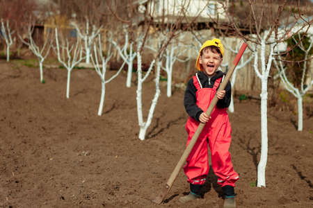 A child with a garden tool in a vegetable garden next to garden trees works and helps plant trees and vegetables in the countrysideの写真素材