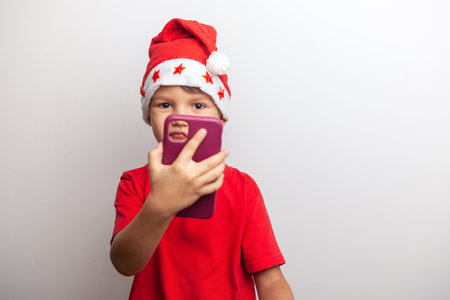 A young boy wearing a Santa hat and a red shirt takes a selfie with a purple smartphone. He is...