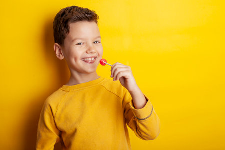 A young boy wearing a yellow sweatshirt against a bright yellow background playfully holds a red lollipop, sticking his tongue out in anticipation. The scene exudes energy and joy with vibrant tones.の写真素材